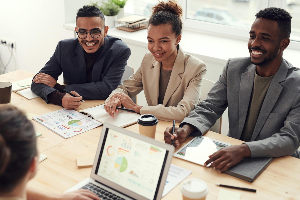 Group of young professionals in a business meeting, smiling and collaborating with charts, graphs, and laptops, representing teamwork and leadership skills gained through a Master’s in Management.