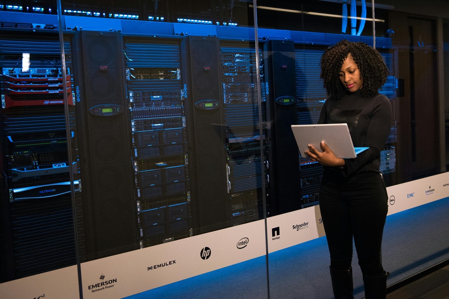 A person stands in front of a large glass window overlooking a server room filled with racks of networking equipment and servers, holding a laptop.