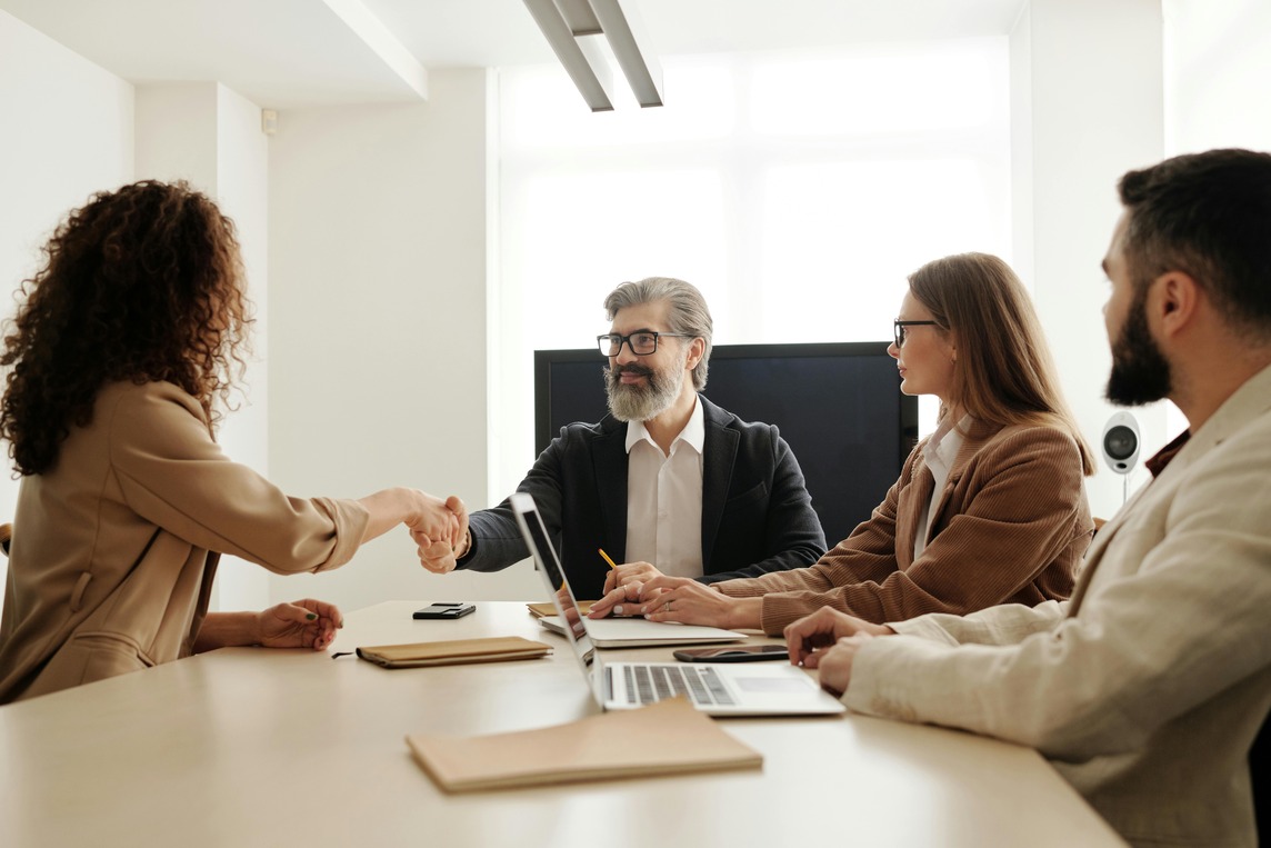 A candidate shaking hands with an interviewer in a professional setting, symbolizing career opportunities with a master’s in human resource management. Three other professionals observe and take notes during the meeting.