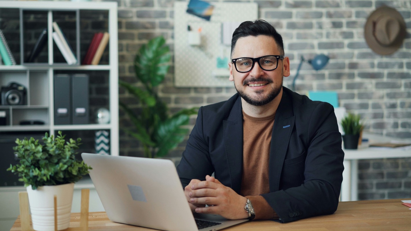 Confident professional sitting at a desk with a laptop, smiling at the camera in a modern office setting—representing a career in project management.