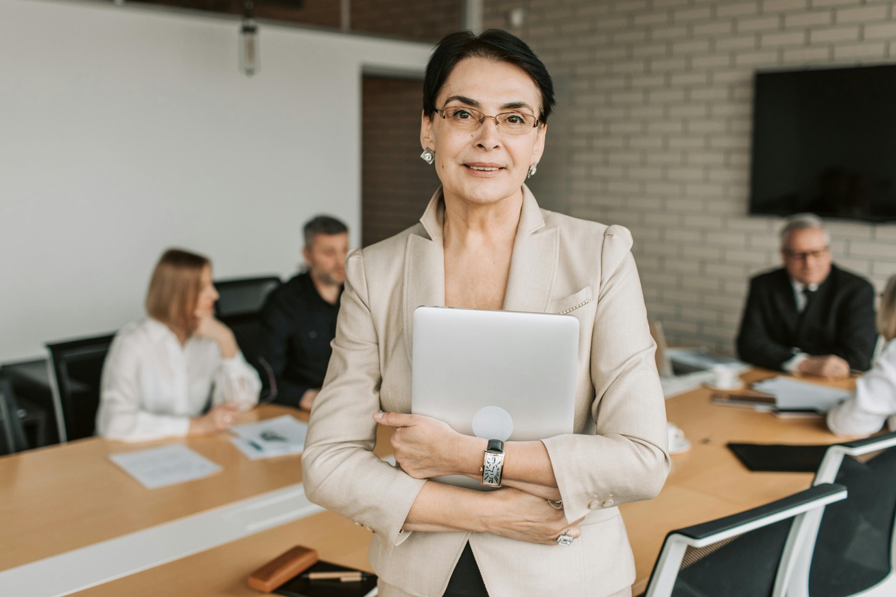Confident businesswoman in a beige suit holding a laptop, standing at the head of a conference table with four colleagues seated in discussion in the background.