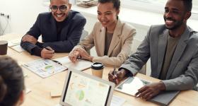 Group of young professionals in a business meeting, smiling and collaborating with charts, graphs, and laptops, representing teamwork and leadership skills gained through a Master’s in Management.