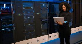 A person stands in front of a large glass window overlooking a server room filled with racks of networking equipment and servers, holding a laptop.