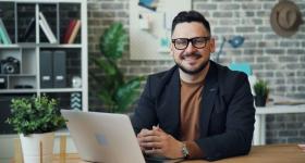 Confident professional sitting at a desk with a laptop, smiling at the camera in a modern office setting—representing a career in project management.