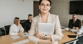 Confident businesswoman in a beige suit holding a laptop, standing at the head of a conference table with four colleagues seated in discussion in the background.